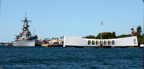 A battleship docked near a white memorial structure on the water, with clear blue skies and palm trees in the background.