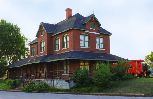 Historic brick train station with a steep roof, surrounded by greenery and a vintage train car nearby.