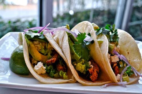 Three tacos filled with chicken, avocado, lettuce, and cilantro, served with lime wedges on a white plate.