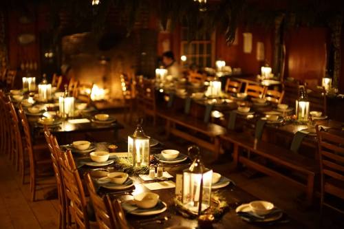 A warmly lit dining room with long tables set for a meal, featuring lanterns and a cozy fireplace in the background.