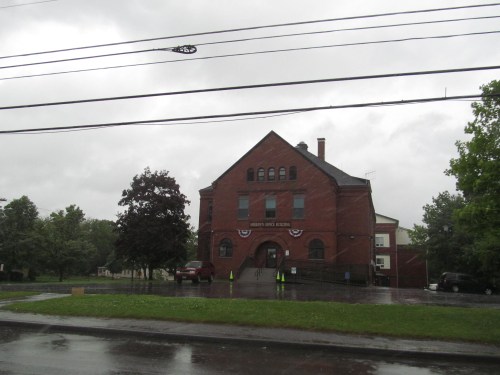 A brick building with a steep roof, surrounded by trees, on a rainy day with power lines overhead.