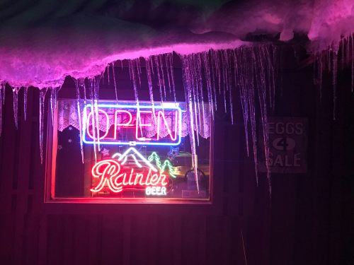 A brightly lit "OPEN" sign and "Rainier Beer" neon sign, with icicles hanging from a snowy roof above.