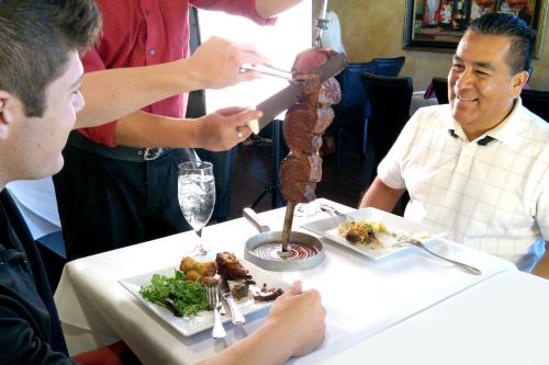 A server carves meat from a skewer for two diners at a restaurant, with plates of food and drinks on the table.