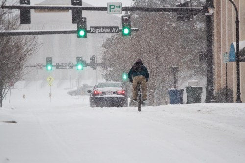 A cyclist rides through a snowy street with green traffic lights, while a car drives nearby in heavy snowfall.