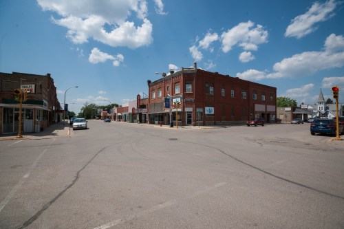 A quiet street scene featuring brick buildings, a clear sky, and a few parked cars in a small town.