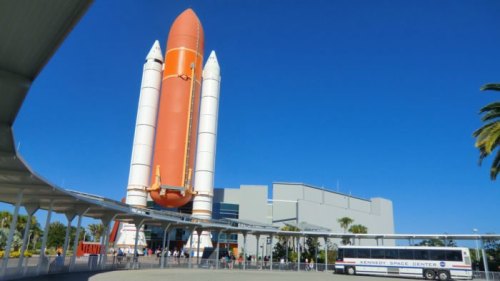A large orange rocket stands tall at the Kennedy Space Center under a clear blue sky, with visitors nearby.