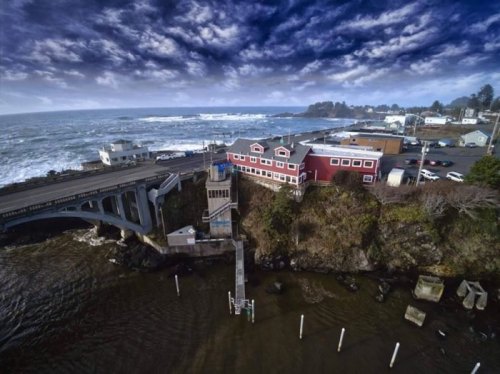 Aerial view of a coastal town with a red building, bridge, and waves crashing against the shore under a cloudy sky.