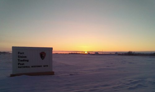 Sunrise over snowy landscape with a sign for Fort Union Trading Post National Historic Site in the foreground.