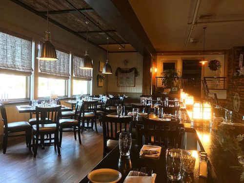 A cozy restaurant interior with wooden tables, chairs, and soft lighting, featuring large windows and elegant decor.