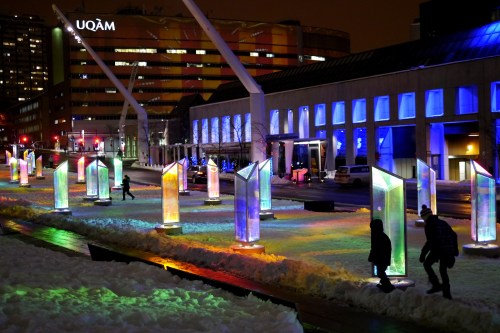 Colorful illuminated ice sculptures line a snowy street at night, with buildings and city lights in the background.