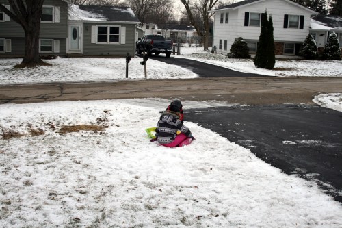 A child sits on a snowy patch in a suburban neighborhood, playing with a sled near a driveway and houses.