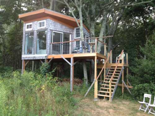 A modern treehouse with large windows, elevated on stilts, surrounded by trees and greenery, featuring a wooden deck and stairs.