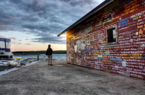 A person stands on a dock beside a colorful mural on a building, with water and clouds in the background.