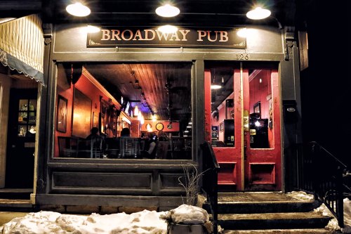 A nighttime view of Broadway Pub, featuring red doors and warm interior lighting, with snow on the ground outside.