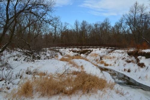 A snowy landscape with a creek and a bridge in the background, surrounded by bare trees and dry grass.
