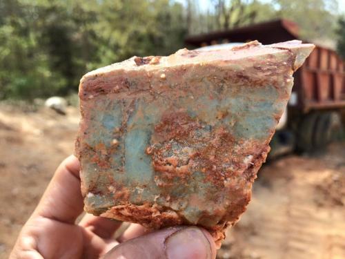 A hand holding a rough, multicolored mineral specimen with green and reddish hues, set against a blurred background.