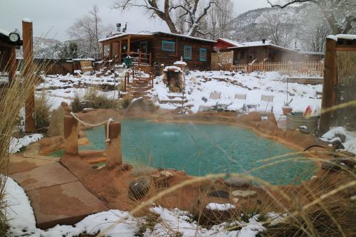 A serene hot spring surrounded by snow, with wooden structures and lounge chairs in a tranquil outdoor setting.