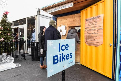 A sign reading "Ice Bikes" in front of a booth with people waiting in winter attire. Christmas decorations are visible nearby.