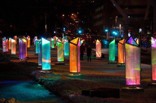Colorful illuminated ice sculptures in a snowy urban setting, with people walking among them at night.