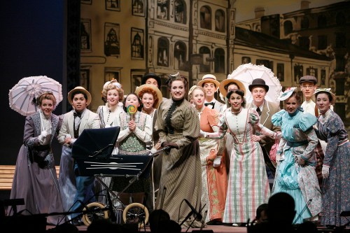 A lively theater cast in period costumes poses together on stage, with a stroller and vintage props in a colorful setting.