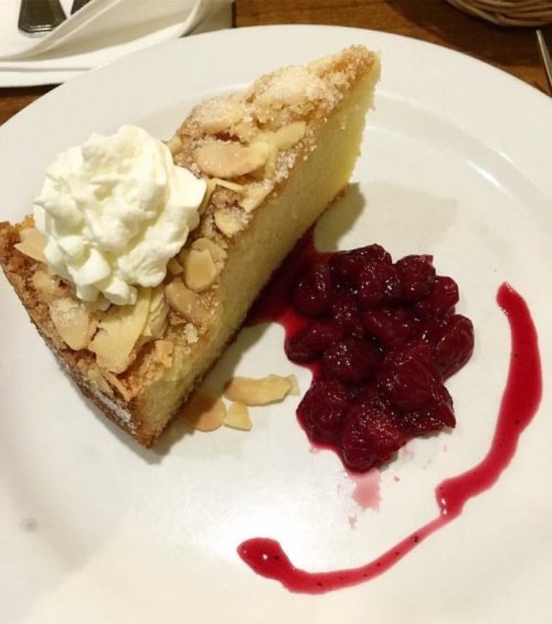 A slice of almond cake topped with whipped cream, served with a side of raspberry sauce and berries on a white plate.