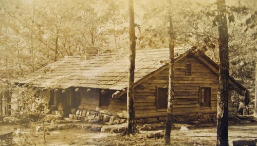 A vintage photo of a rustic log cabin surrounded by trees in a natural setting.