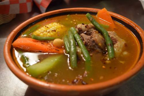 A bowl of hearty soup with meat, corn, carrots, green beans, and spices, served in a traditional clay dish.