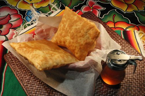 Two golden, crispy pastries on parchment paper, accompanied by a small jar of honey, set against a colorful floral background.