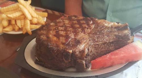 A large grilled steak served on a plate with a slice of watermelon and a side of crispy French fries.