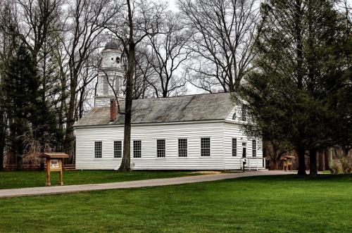 A historic white building surrounded by trees, with a bell tower visible in the background and a grassy area in front.
