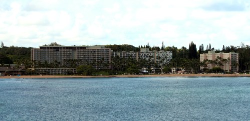 A coastal view of a modern hotel complex surrounded by palm trees and lush greenery, with calm waters in the foreground.