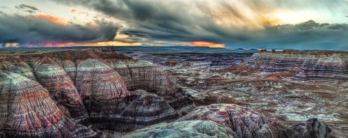 A panoramic view of colorful rock formations under a dramatic sky at sunset in a desert landscape.