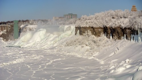 Frozen Niagara Falls with icy cliffs and snow-covered trees under a clear blue sky.