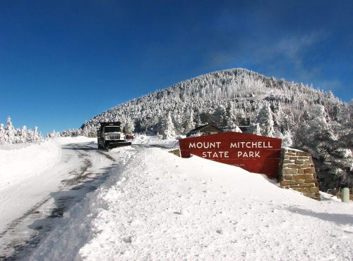 Snow-covered landscape at Mount Mitchell State Park, with a sign and a vehicle on a snowy road under a clear blue sky.