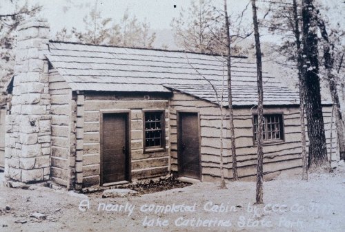 A nearly completed log cabin in Lake Catherine State Park, surrounded by trees and featuring a stone chimney.