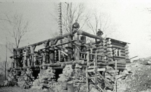 Black and white photo of a log cabin under construction, with two men standing on the structure. Trees in the background.