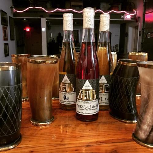 Three bottles of craft beverages surrounded by decorative glasses on a wooden table in a cozy setting.