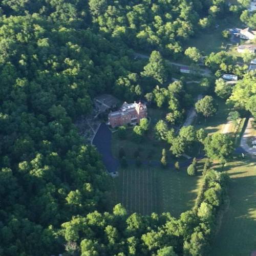 Aerial view of a large, historic building surrounded by dense green trees and open fields.