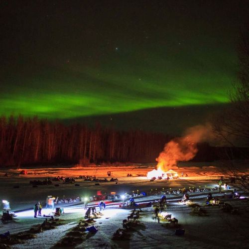 A snowy landscape at night with people gathered around a fire, under vibrant northern lights in the sky.