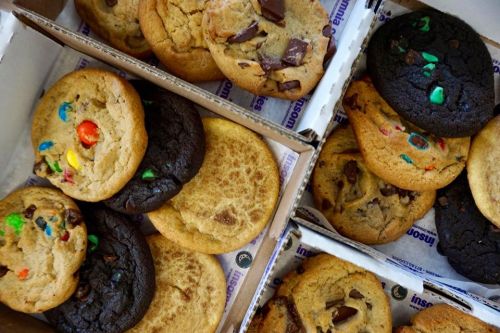 A variety of cookies in two boxes, including chocolate chip, M&M, and dark chocolate cookies.