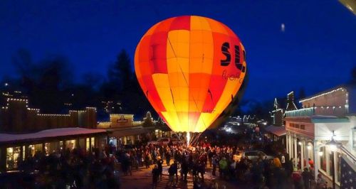 A vibrant hot air balloon glows against a night sky, surrounded by a crowd and festive lights in a lively town.