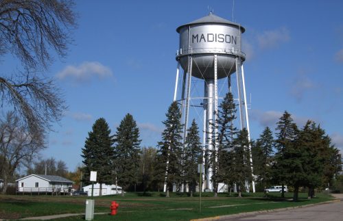 A tall silver water tower labeled "MADISON" stands among green trees under a blue sky.