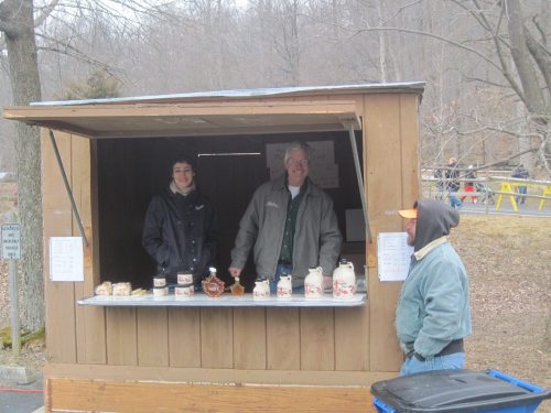 Two vendors smile from a wooden stand, offering jars and baked goods, with a customer in a hoodie nearby.