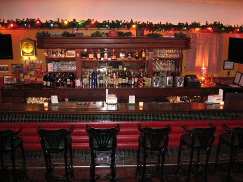 A cozy bar with a wooden counter, shelves of liquor, and festive decorations, including lights and greenery.
