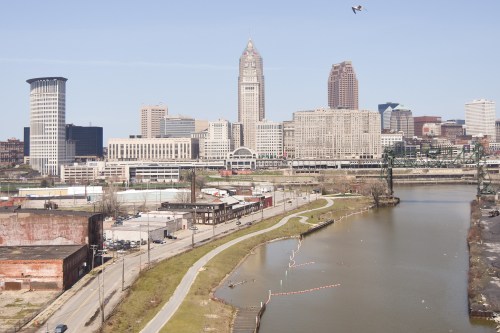 A city skyline featuring tall buildings alongside a river, with a clear blue sky and a bird flying overhead.