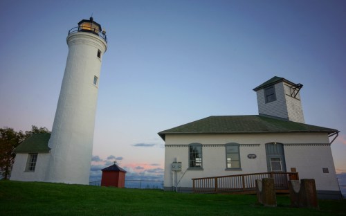 A white lighthouse beside a small building, set against a twilight sky with soft clouds. Green grass in the foreground.