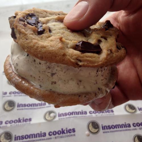 A hand holding a chocolate chip cookie ice cream sandwich, with melting ice cream and a branded paper underneath.