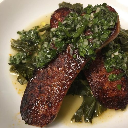 Two grilled sausages topped with a green herb sauce, served on a bed of sautéed greens in a white bowl.