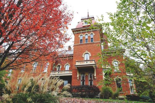 A historic brick house with a tower, surrounded by colorful autumn foliage and tall grasses.