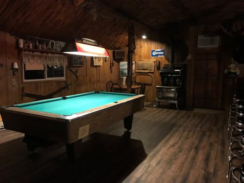 A cozy room with a green pool table, wooden walls, and rustic decor, featuring a stove and bar stools in the background.
